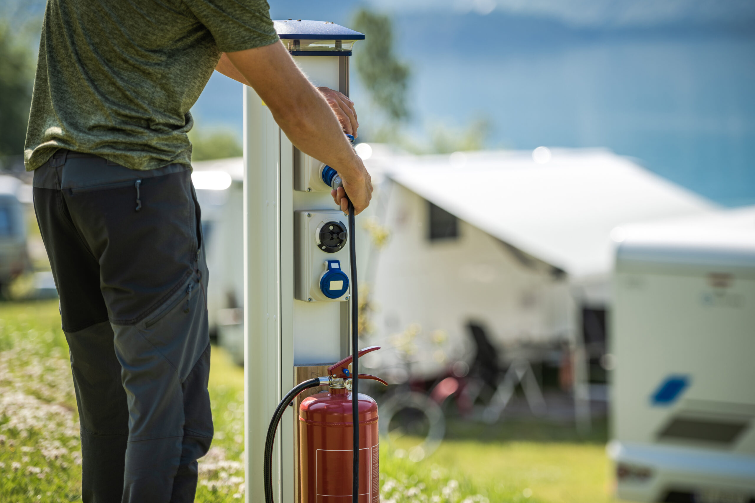 A person stands outdoors beside a camper van, connecting a power cable to an electric hook-up station next to a red fire extinguisher at a campsite with grass and distant water in the background.