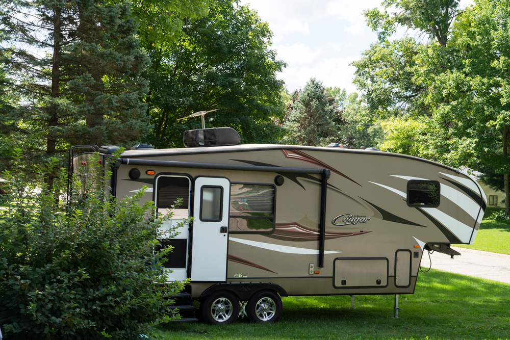A beige fifth-wheel RV labeled "Cougar" is parked on grass near trees and bushes, with sunlight filtering through the leafy green branches.