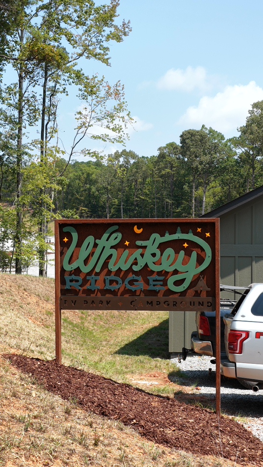 A rustic wooden sign with "Whiskey Ridge" painted in green and white stands by a gravel lot with a white SUV, trees, and a gray building in the background under a blue sky.