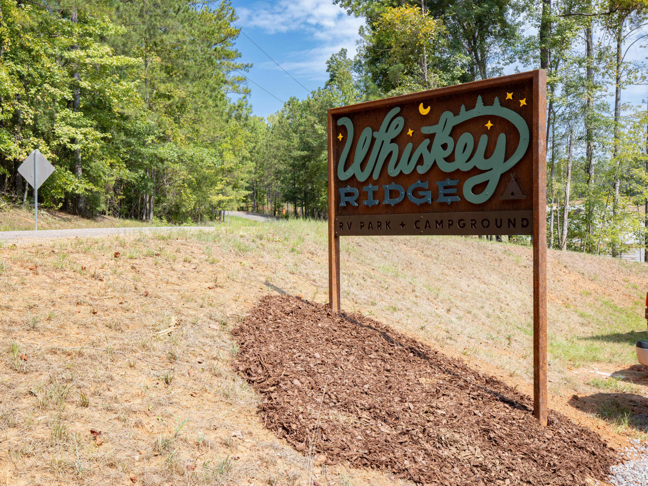 A large wooden sign reads “Whiskey Ridge RV Park & Campground” on a grassy slope beside a road, surrounded by trees.