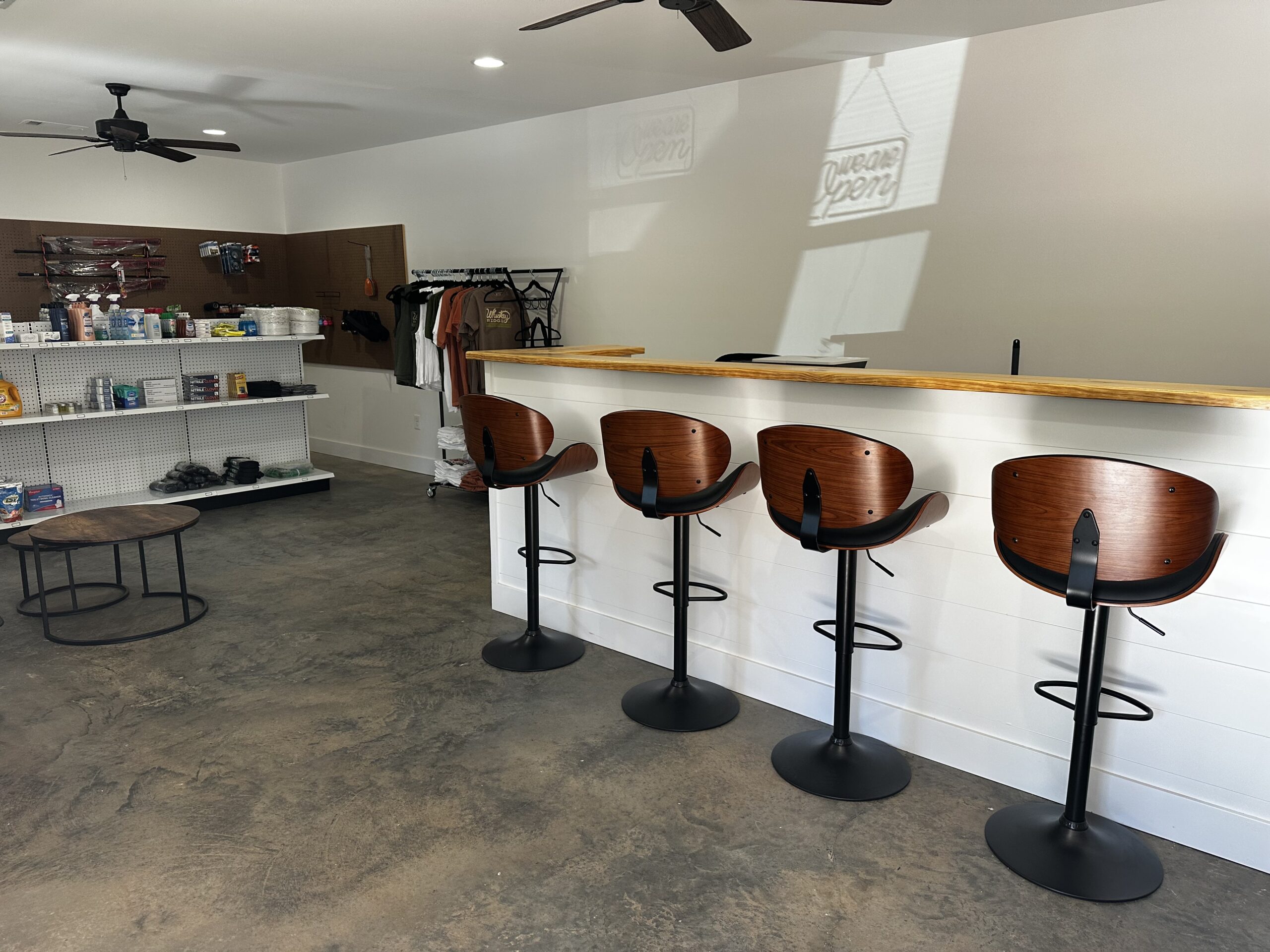 A modern retail space with four wooden bar stools at a counter, shelves with merchandise, and a clothing rack against a white wall.