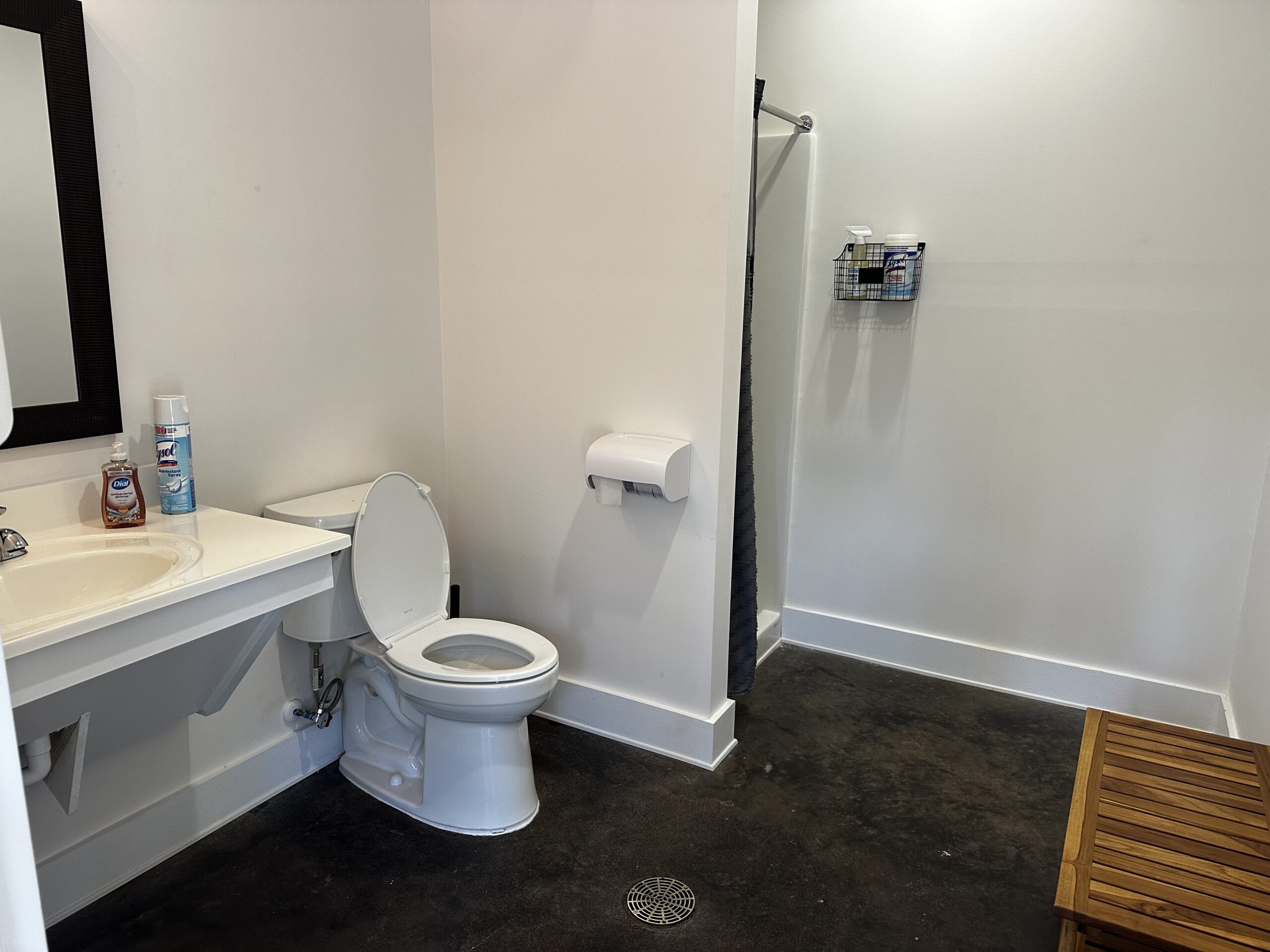 Accessible bathroom with a sink, toilet, wall-mounted toilet paper, open shower area with toiletries, and a wooden bench on a dark floor.