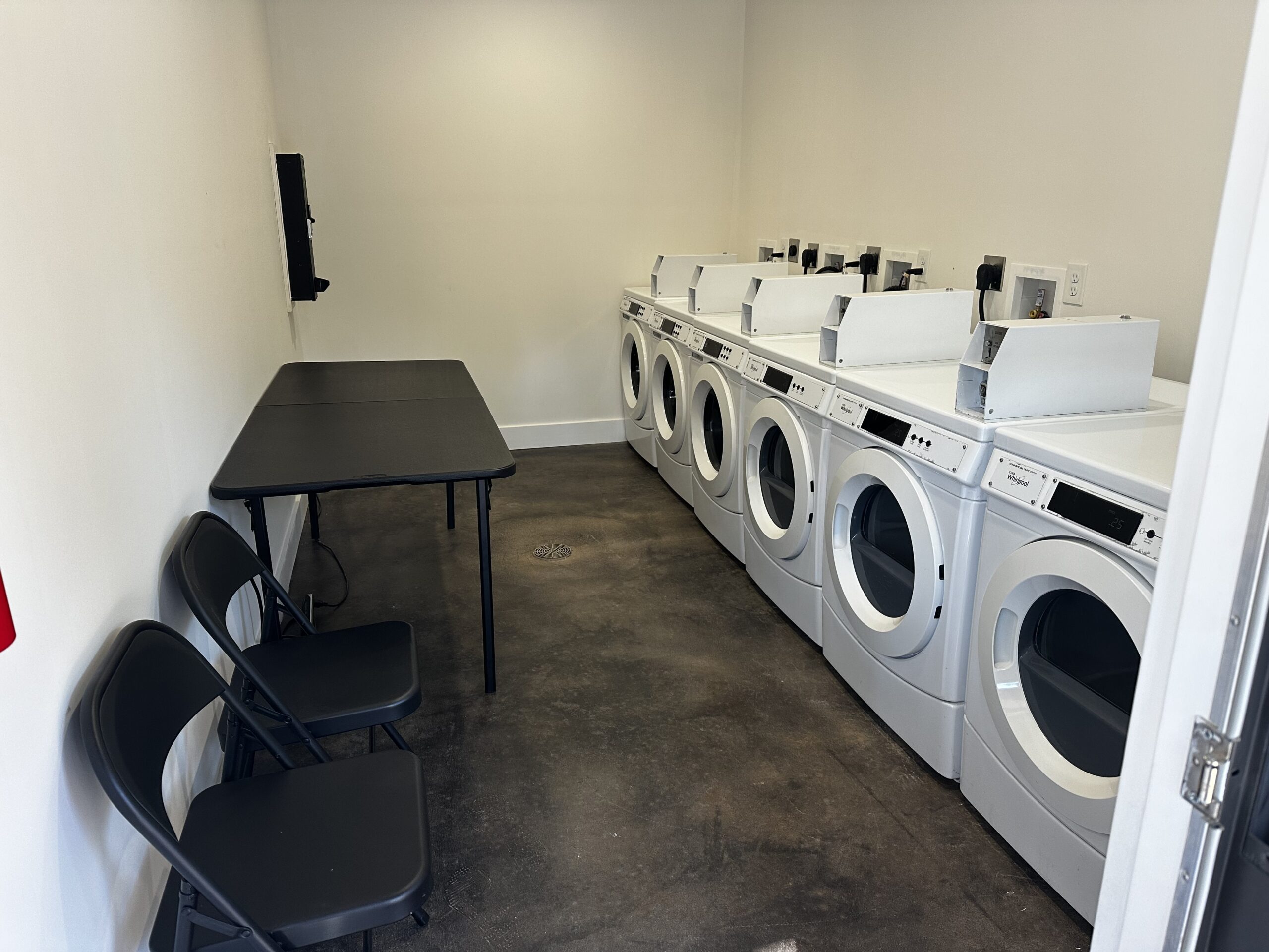 A small laundry room with six front-loading washing machines, a black folding table, and two black folding chairs against the wall.