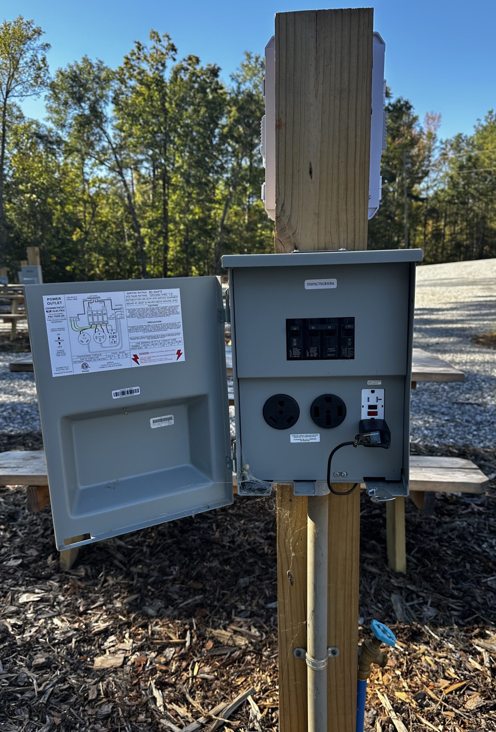 An open outdoor electrical panel mounted on a wooden post, showing circuit breakers, outlets, a GFCI outlet, and wiring diagrams inside the door.